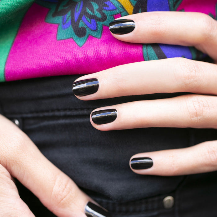 A close-up of a hand with neatly manicured nails painted in Licorice by Manucurist, resting on a hip. The person wears black pants and a vibrant pink and green patterned top.