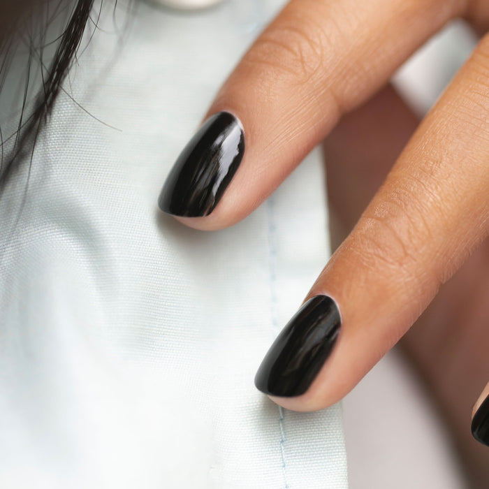 A close-up of a hand with neatly manicured nails painted in Manucurist Licorice black polish, resting on light blue fabric.