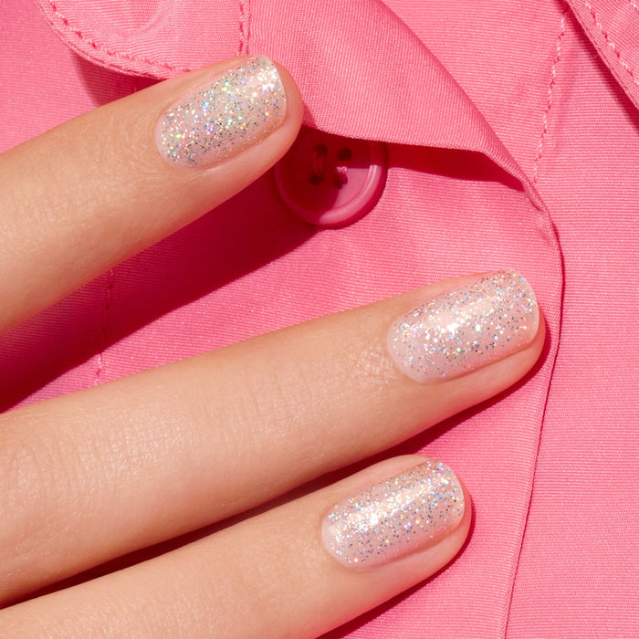 Three fingers with short nails painted in Manucurist's Mermaid sheer holographic glitter polish, resting on a bright pink shirt with a visible pink button.