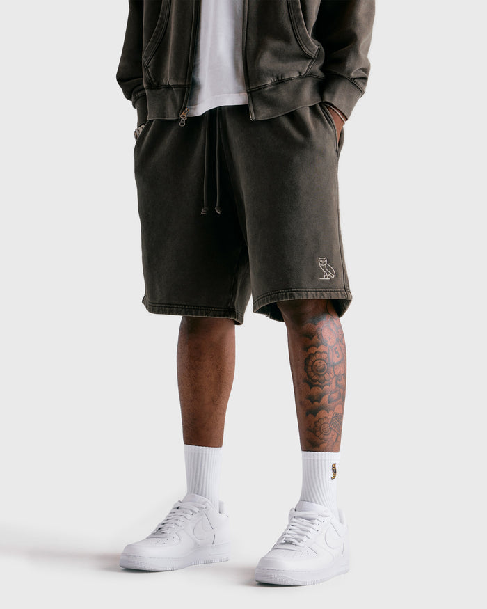 Deep olive sweatshorts with drawstring waist and side pockets, styled with black jacket and white sneakers.