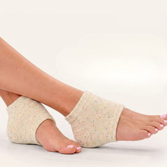 A person wears LONDONTOWN Pedi Perfect Lightweight Heel Socks in beige speckle, designed for heel therapy, with bare heels and toes visible while sitting cross-legged against a plain background.
