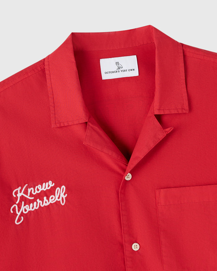 Red camp shirt with white embroidered text on chest and notched collar, displayed on white background.