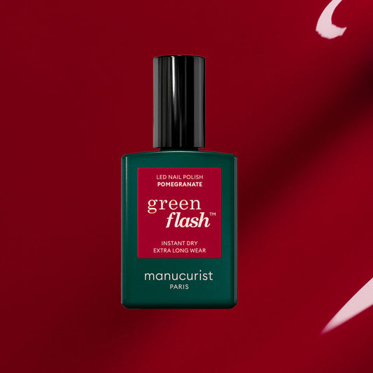 A bottle of Manucurist’s Pomegranate Green Flash gel polish, a vibrant red shade, stands upright on a red background with white accents. The dark green bottle with a red label and black cap adds elegance to any nail polish collection.
