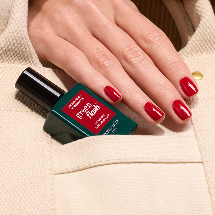 A hand with glossy Manucurist Pomegranate gel polish holds a bottle of Green Flash by Manucurist, partially tucked into the pocket of a beige textured garment.