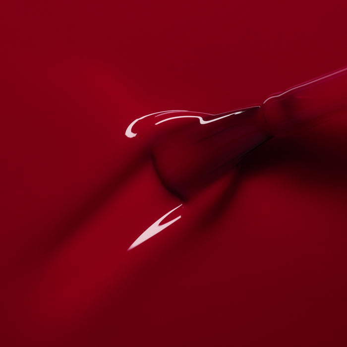 A close-up shows a dropper releasing Manucurist’s Pomegranate gel polish—a glossy raspberry red—onto a smooth red surface, forming a shiny, flowing puddle.