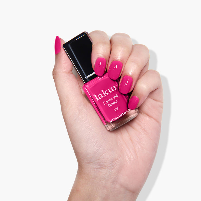 A hand with long, manicured nails is holding a bottle of pink nail polish labeled "Queen of Hearts by LONDONTOWN." The vegan, cruelty-free polish is a deep fuchsia pink crème, and the nails are painted in the same vibrant shade.