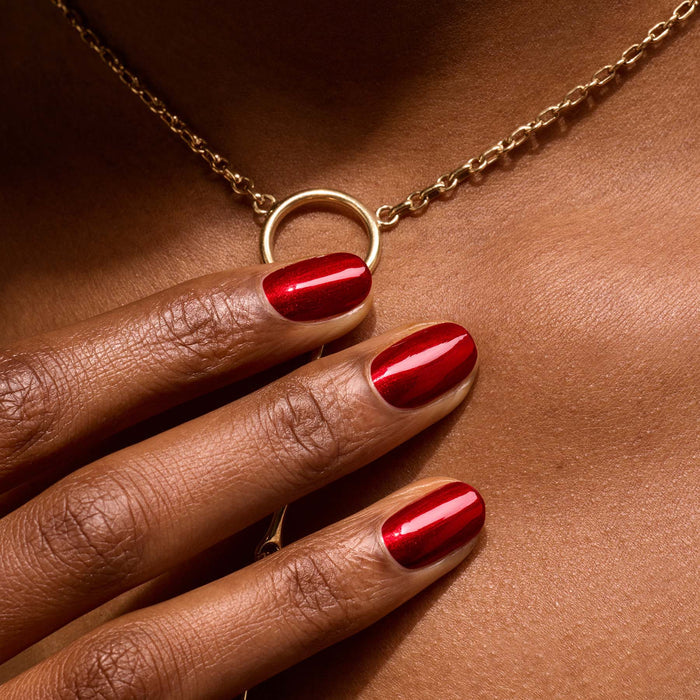 A close-up of a hand with Manucurist Red Hibiscus nail polish rests on a chest, highlighting a gold necklace with a circular pendant.