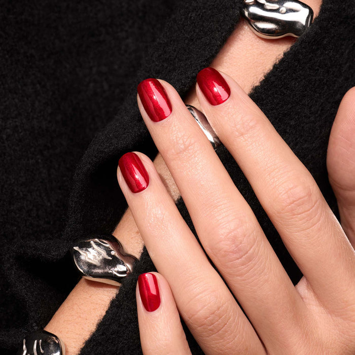 A close-up of a hand with Manucurist’s Red Hibiscus polish, resting on black textured fabric. The look is completed with chunky silver rings and a matching bracelet in smooth, organic shapes.