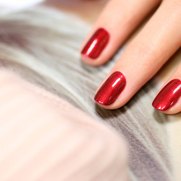 A close-up of a hand with neatly manicured nails painted in Manucurist's Red Hibiscus gel polish, resting on a soft, blurred background.