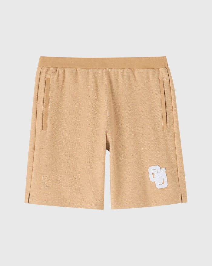 Camel-colored sweatshorts with elastic waistband, side pockets, and white logo patch on leg.