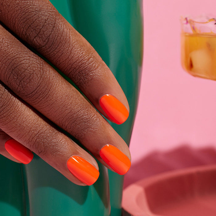A hand with neatly manicured nails painted in Manucurist's Sunset, a vibrant orange polish, rests on a glossy green surface with a pink background and a drink with ice partially visible.