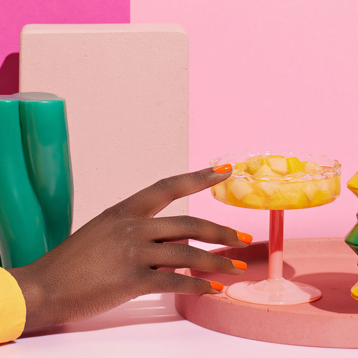 A hand with Manucurist's Sunset vibrant orange nail polish reaches for a pink glass dish of fruit salad, set among colorful geometric objects and a pink background for a modern, lively vibe.