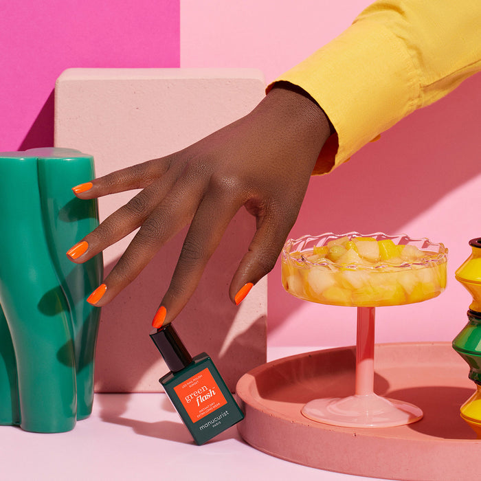 A hand with vibrant orange Manucurist Sunset gel polish holds a matching bottle of Sunset polish, surrounded by colorful decor and a dish of fruit, all set against a pastel pink background.