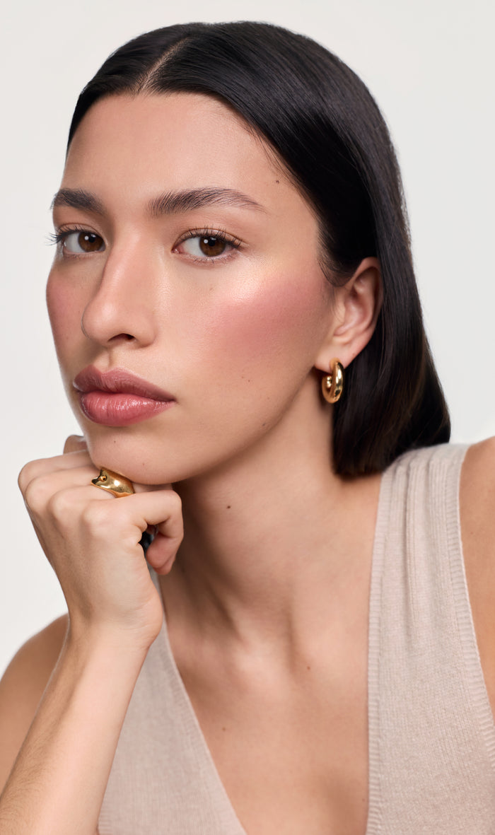 Model wearing makeup with gold earrings against a grey background.