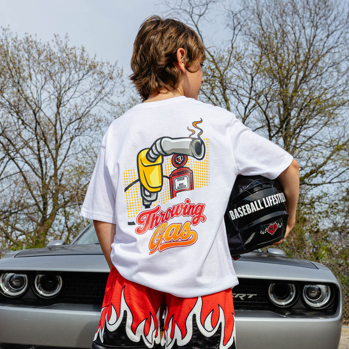 Back view of white youth Throwing Gas tee with colorful gas nozzle and pump graphic, boy holding racing helmet beside silver Dodge Challenger.