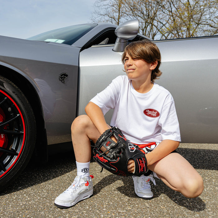Young boy crouching beside silver Dodge SRT Challenger, wearing white Throwing Gas tee with red BL101 chest oval and holding a baseball glove.