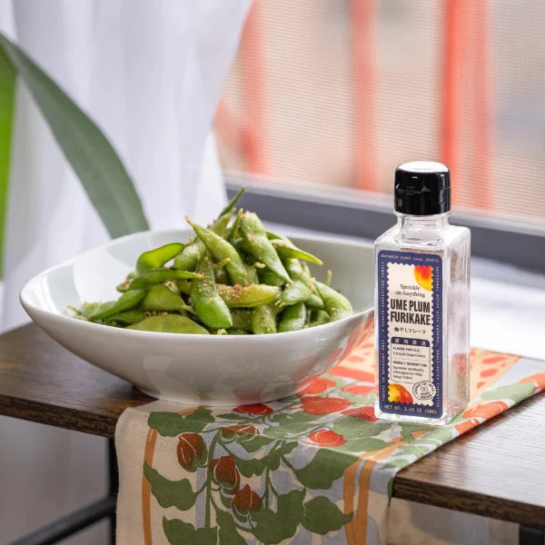 A bottle of One Plum Fumikake seasoning stands next to a white bowl filled with edamame on a wooden table with a floral table runner, set in a bright kitchen environment.